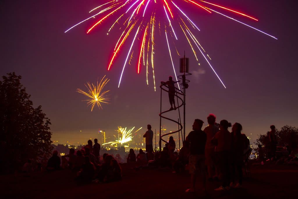 People are silhouetted by the glow of red exploding fireworks behind them — beyond the hill where they're standing, it's obvious more are going off in the distance.