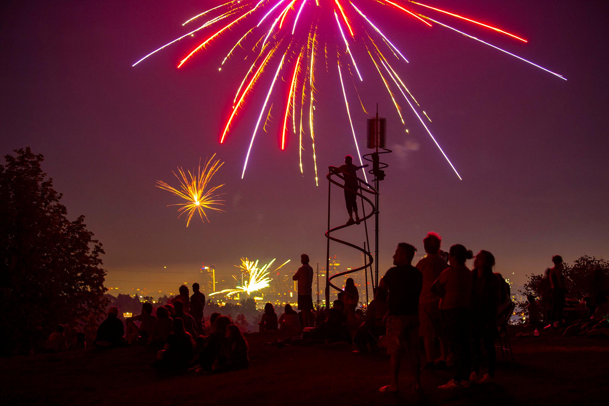People are silhouetted by the glow of red exploding fireworks behind them — beyond the hill where they're standing, it's obvious more are going off in the distance.