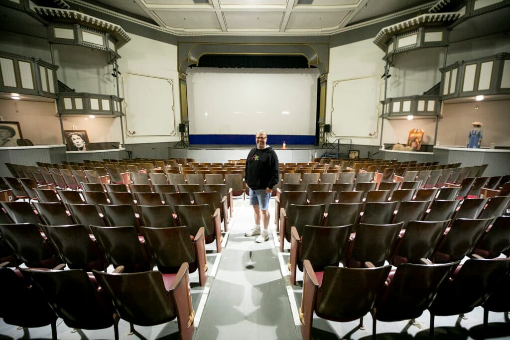 A man in shorts and a hoodie stands in the aisle of an old theater, surrounded by rows of empty chairs and with a stage behind him.