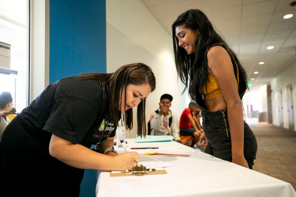 A woman bending over a table signing a paper with another woman standing across the table. A man in the background gives the camera a thumb's up.