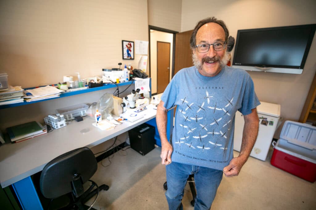 A man with a mustache and excellent side burns wears a goofy smile as he shows off his t-shirt, which is covered with images of mosquitoes.