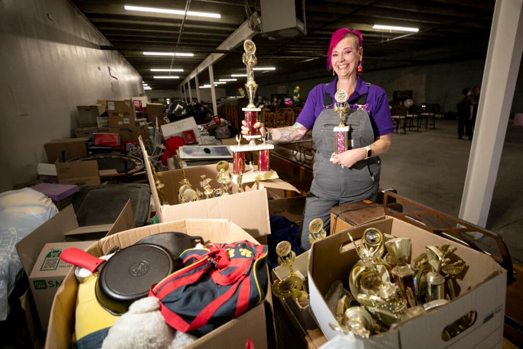 A woman smiles wide, holding old trophies and surrounded by boxes of trophies.