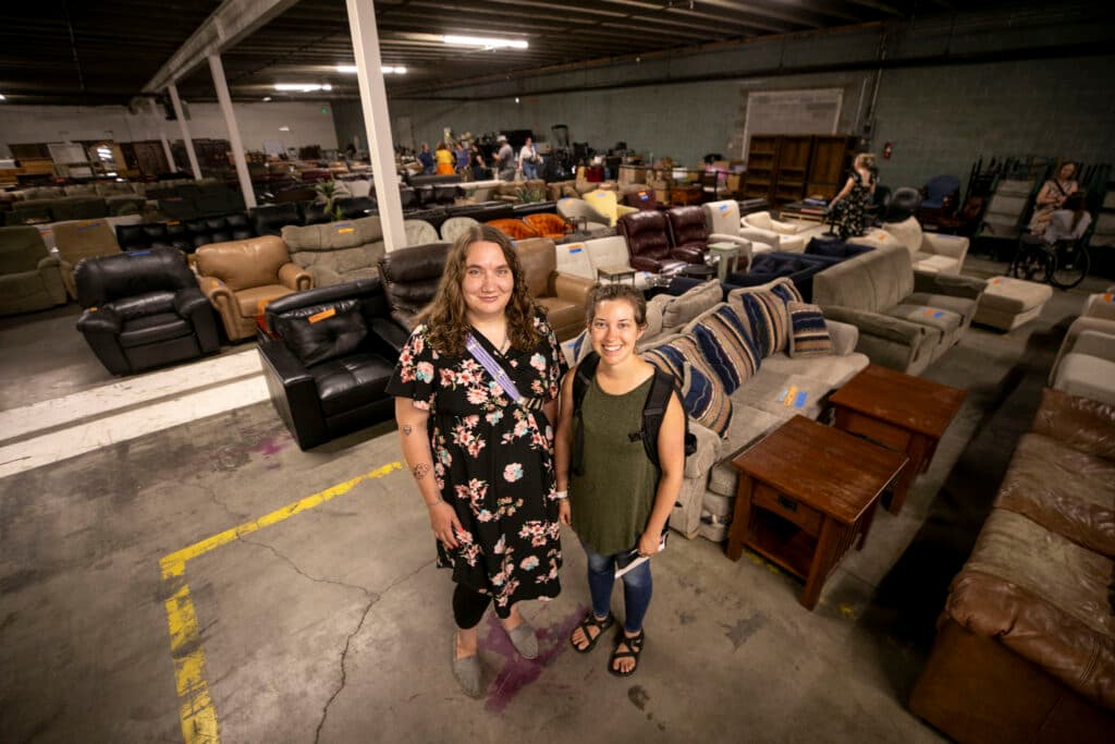Two women stand in a warehouse, surrounded by chairs and couches.