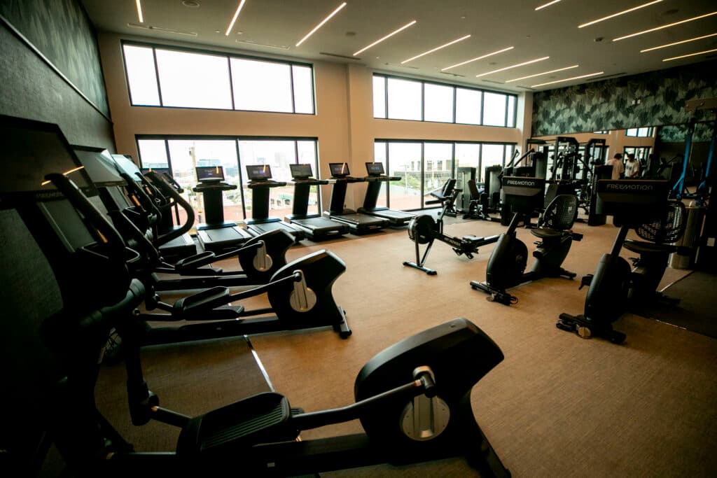 Treadmills and elliptical machines sit on a tan floor, next to a wall of bright windows.