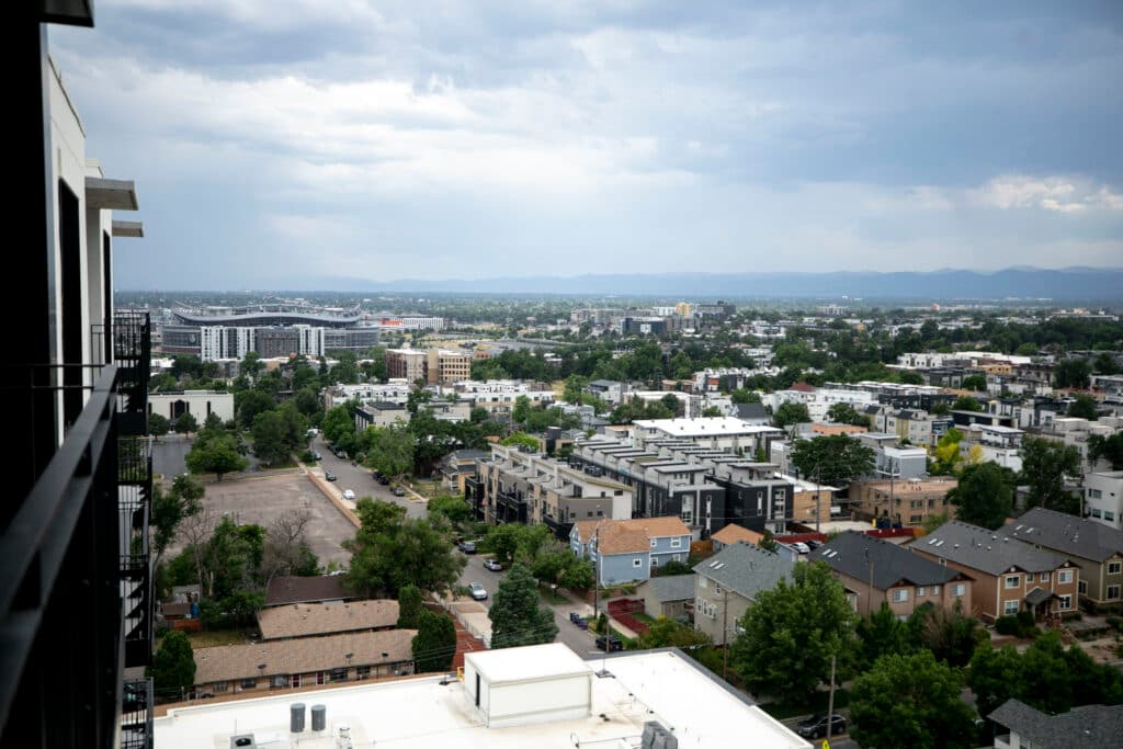 A view over homes in Denver, with Mile High Stadium and the mountains behind it. The sky is blue and cloudy, the trees are green.