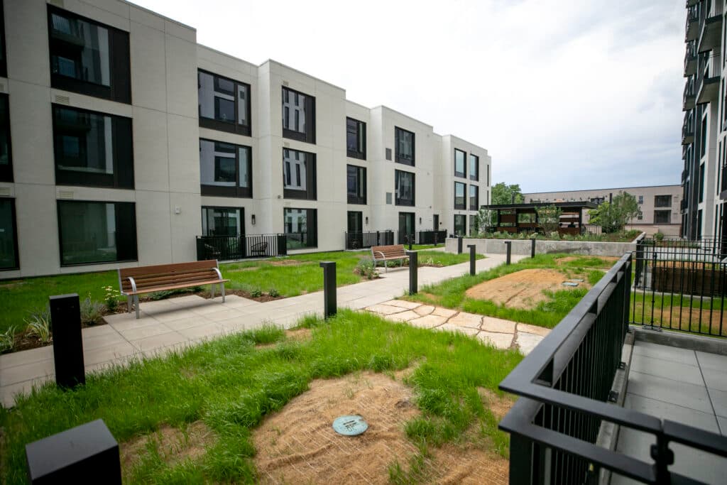 Mostly green grass surrounds a walking path and benches, which are surrounded by apartments.