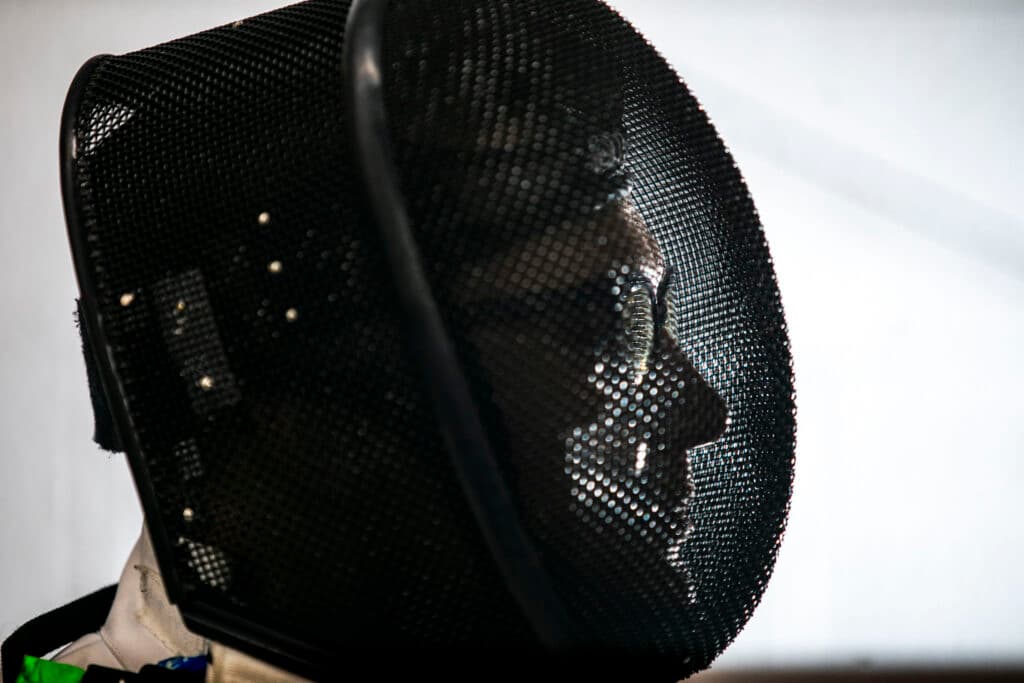 Light illuminates the back side of a fencing mask, revealing the profiled face of a woman behind the protective mesh.