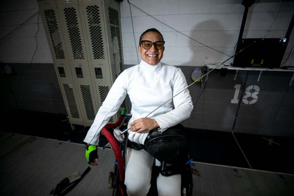 A woman sitting in a wheelchair smiles wide at the camera, holding a fencing sword and wearing a fencing jacket.
