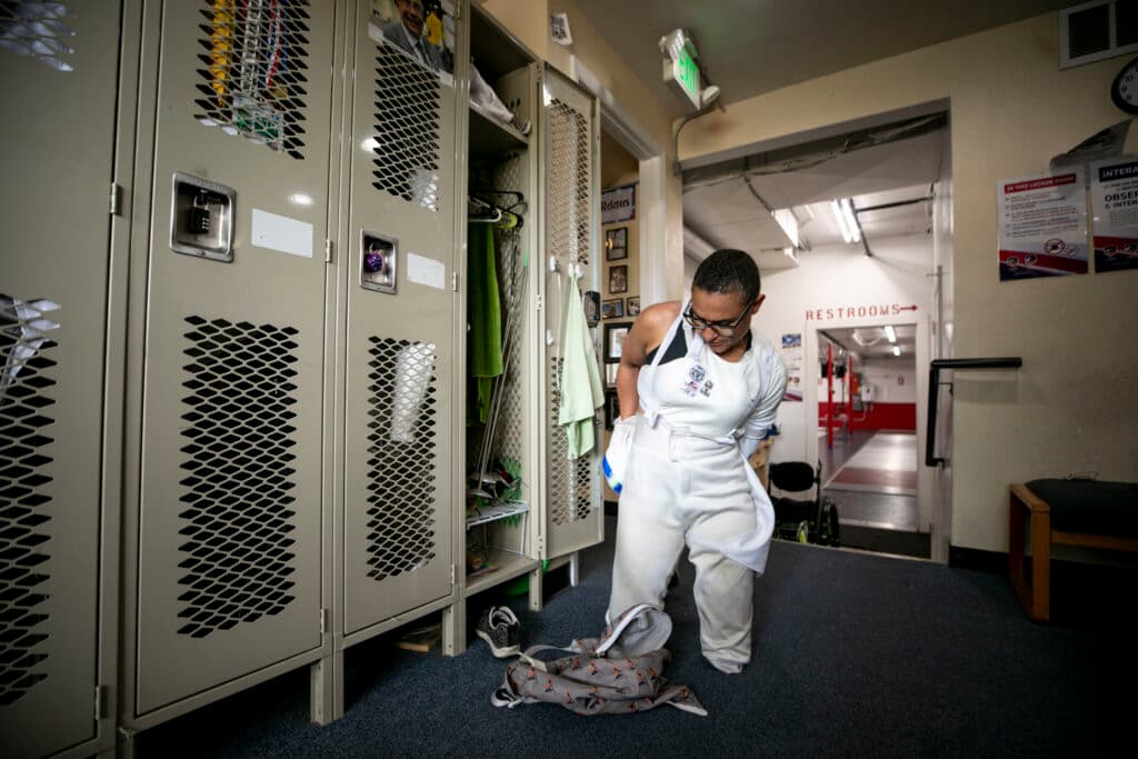 A woman with short hair kneels on the floor as she takes off a fencing vest, in a room lined with lockers.