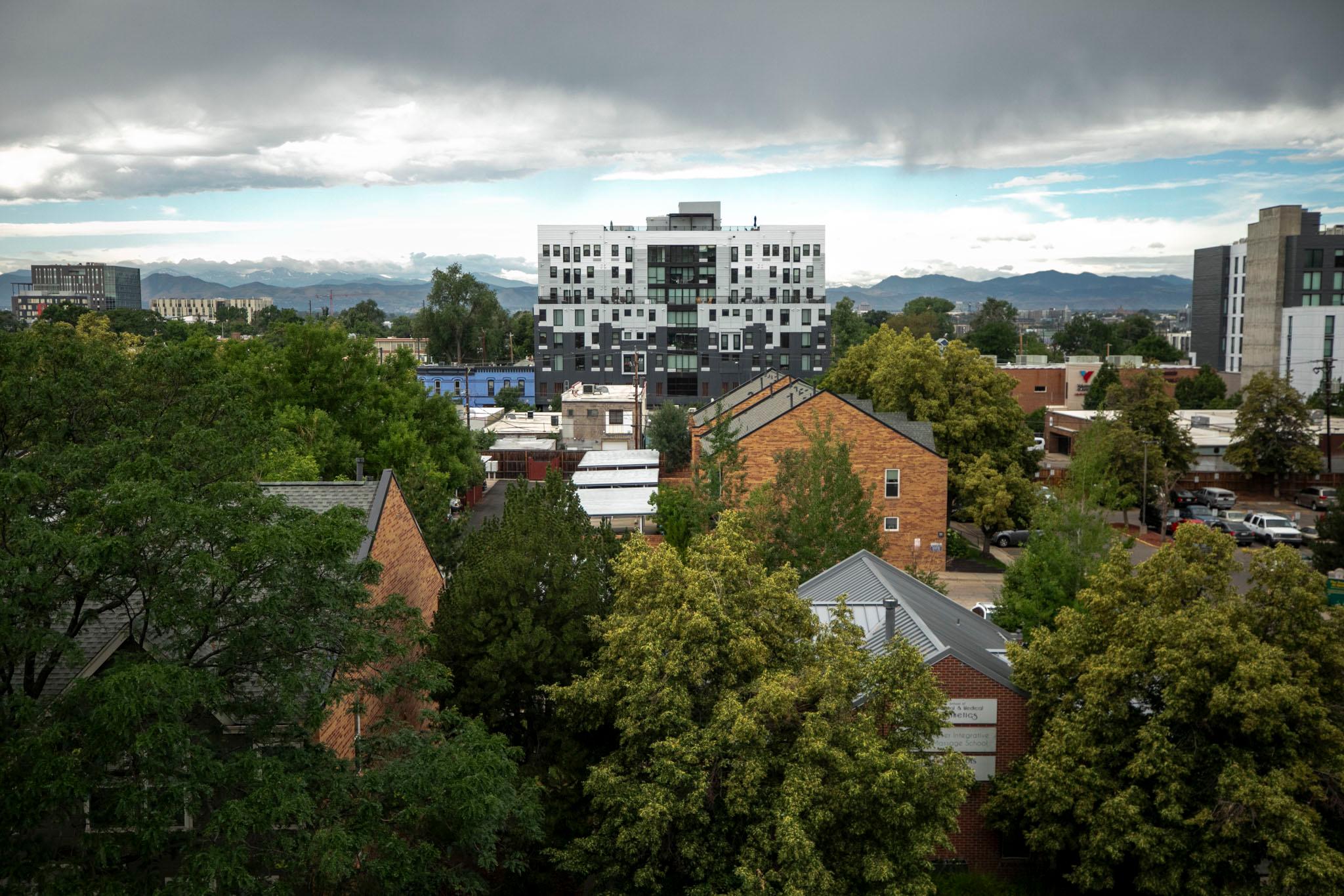 A grey and white building rises from a green neighborhood canopy, with cloudy skies above and the Rockies behind it.