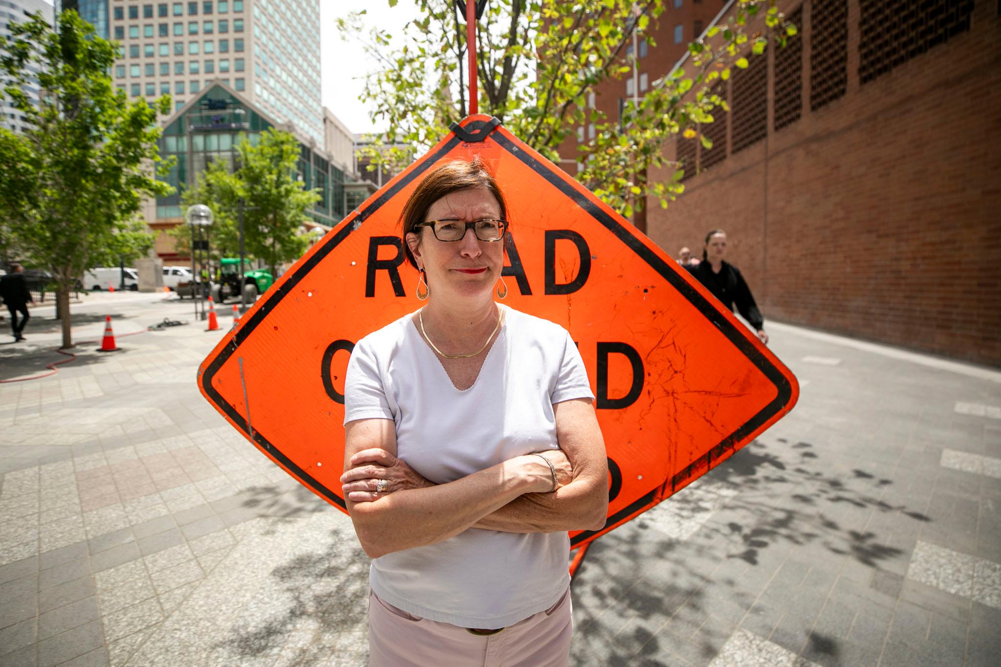 A woman with short hair, a white shirt and glasses stands in front of a construction sign with her arms crossed. The look on her face says "FRUSTRATED!"
