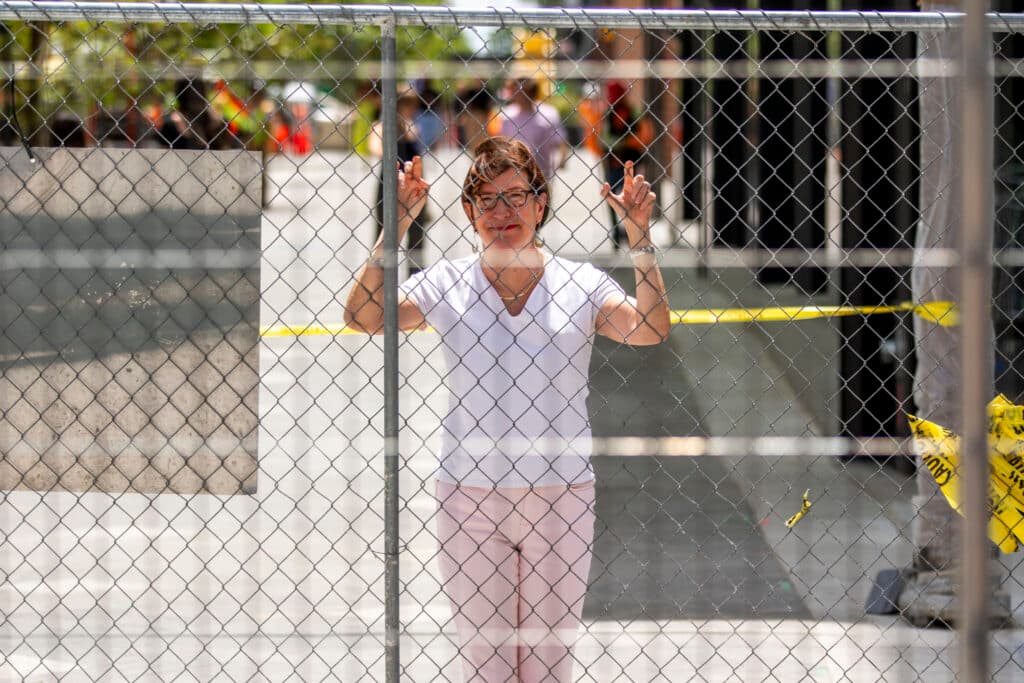 A woman with short hair, a white shirt and glasses leans on a construction fence, looking right into the camera through the chain link.
