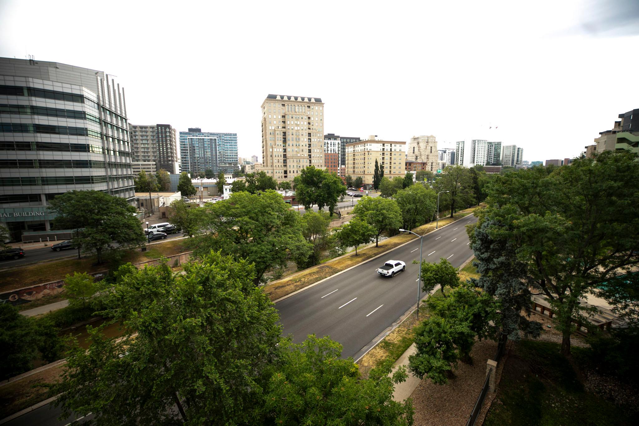 A single white pickup drives on Speer Boulevard, seen from several stories above the road. Apartment buildings rise from the horizon in the background.