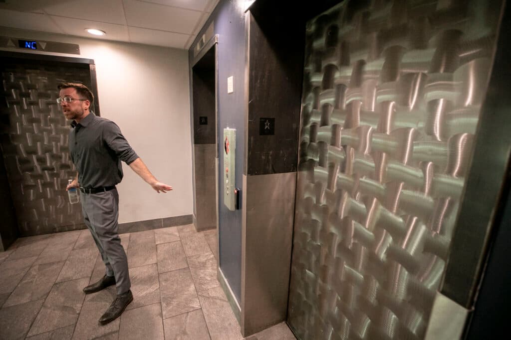 A man in a grey shirt and slacks stands in front of a brushed-metal elevator door. He looks frustrated.