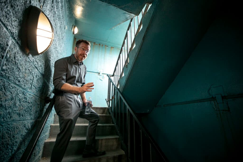 A man in a grey shirt and slacks stands in a turquoise stairwell, speaking with obvious frustration.