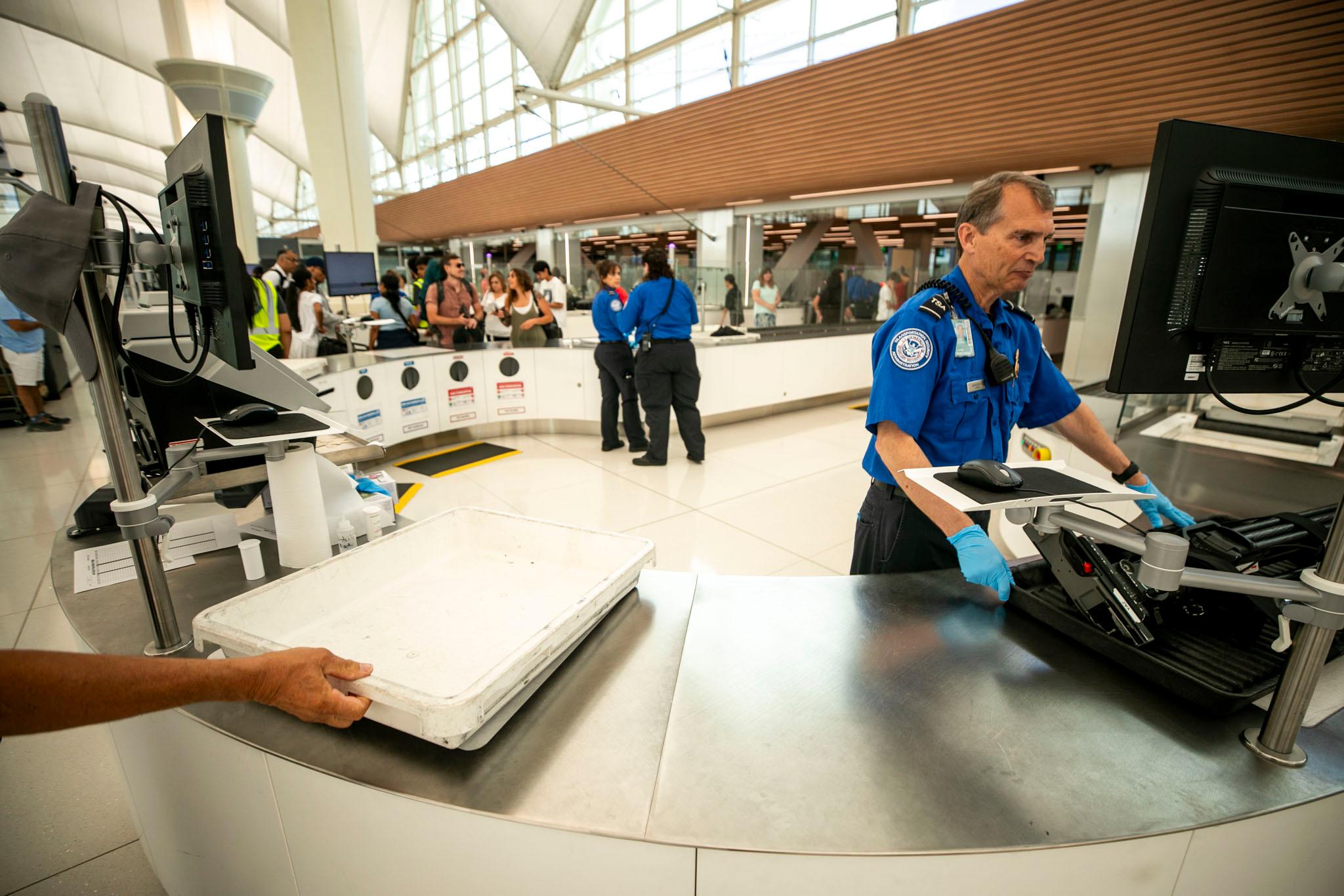 A TSA agent in a blue shirt looks at a screen as a hand drops an empty bin next to him. People go through another line in the background.