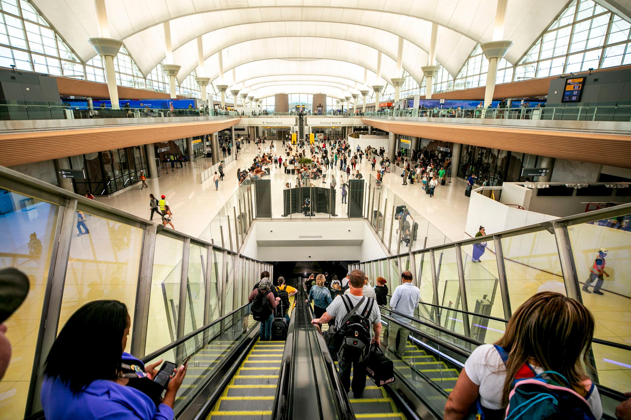 People ride an escalator sloping down through the floor of DIA's main hall. People mill around beyond the hole.
