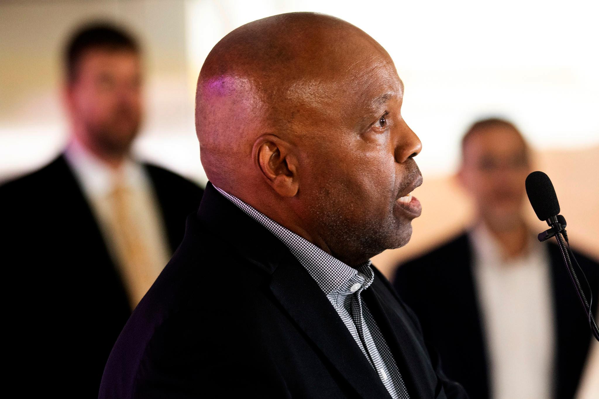 A bald man speaks at a microphone. We can see people in suits out of focus behind him.