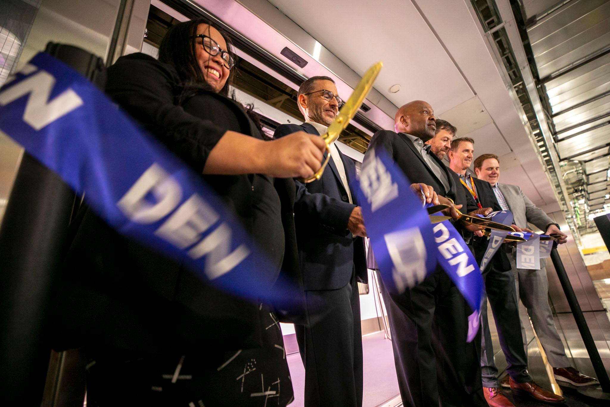 People in suits smile as they use shiny golden scissors to cut a blue ribbon with "DEN" printed on it.
