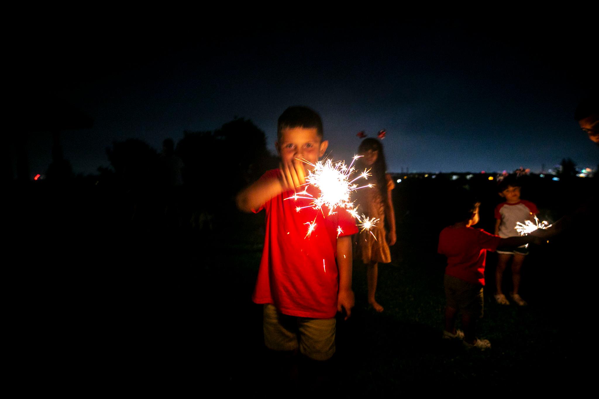 A kid in a red shirt wears a big smile as he swings a glowing sparkler around his head.