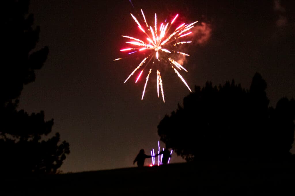 The silhouettes of two people holding hands can be seen in the foreground as the sky behind them is illuminated by bright red fireworks.