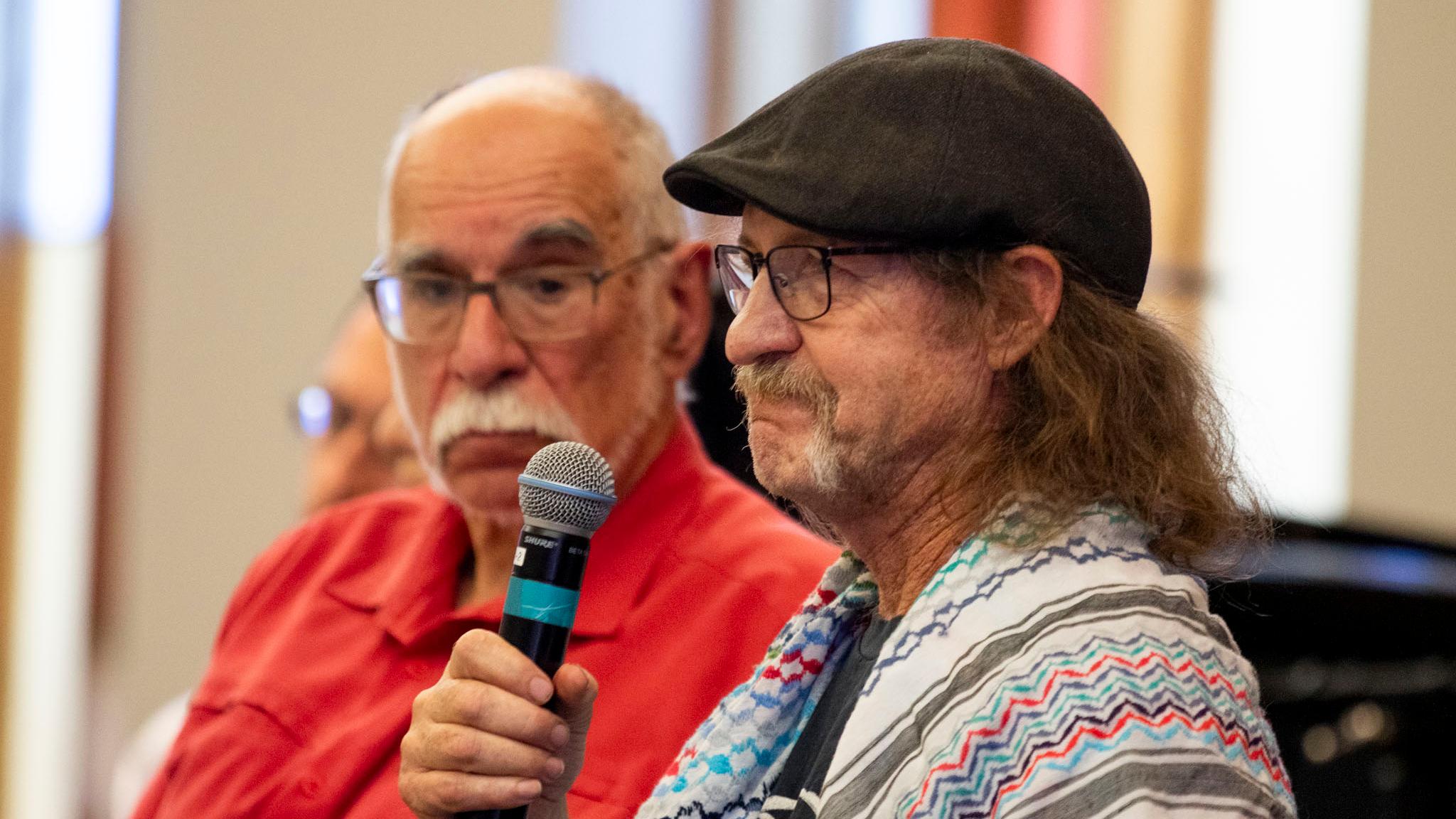 A man in a black cap and a colorful keffiyeh around his neck purses his lips, emotionally, as he looks past the microphone in his hand.