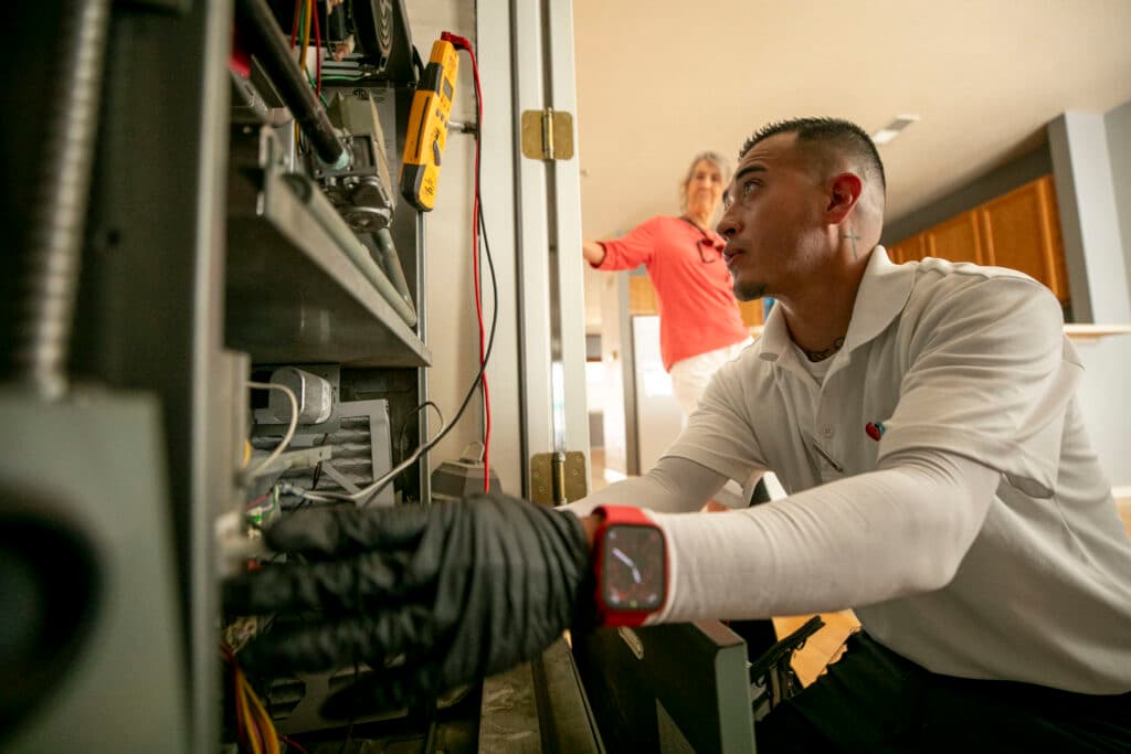 A man with gloved hands reaches into an opening in a wall, overflowing with wires and metal.