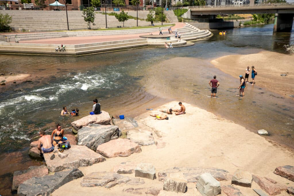 People in bathing suits play in a river, lined by rocks and pavilions.