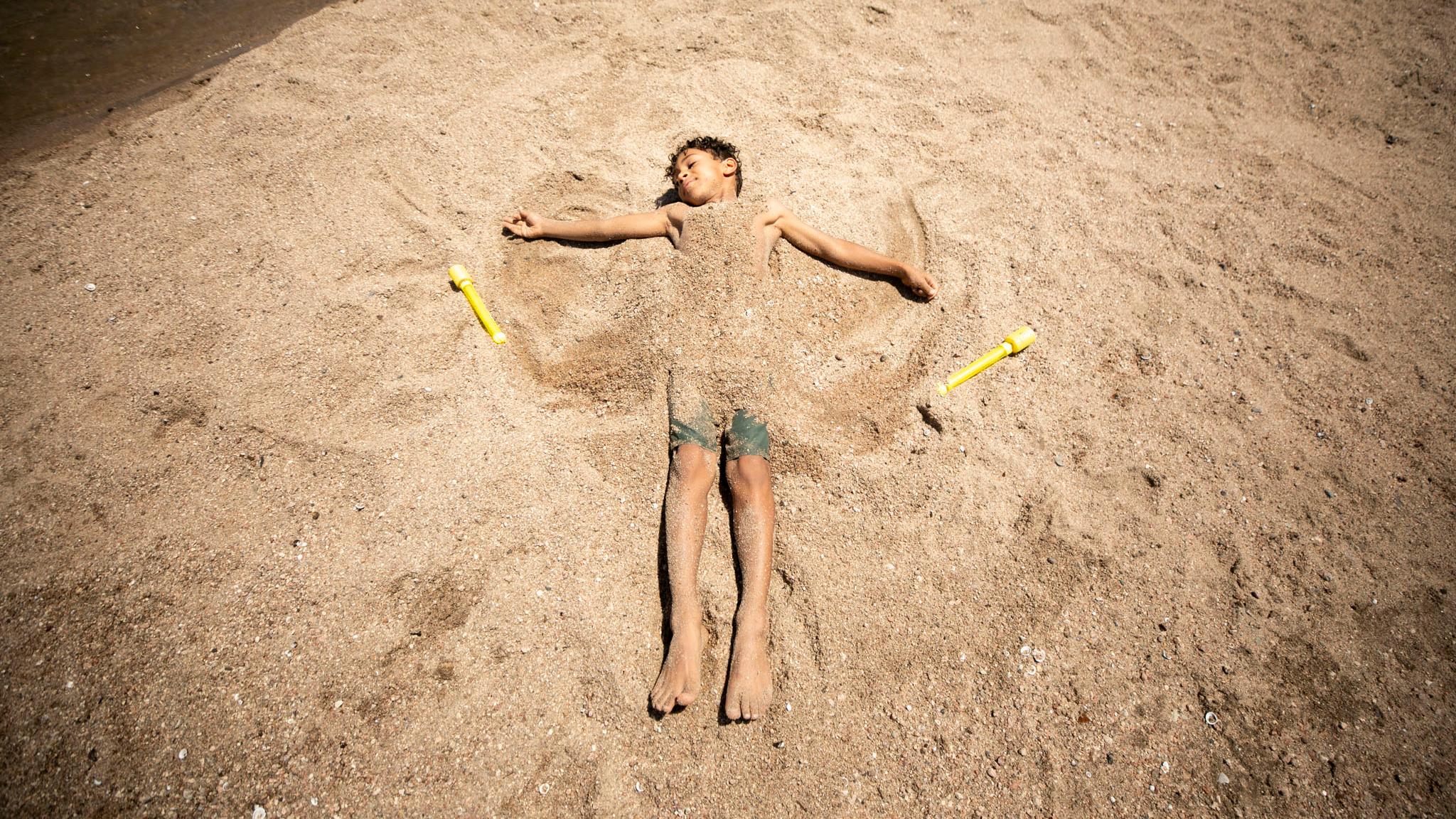 A kid lays in the sand, covered by the sand, with his arms stretched out as if it were winter and he was making a snow angel.
