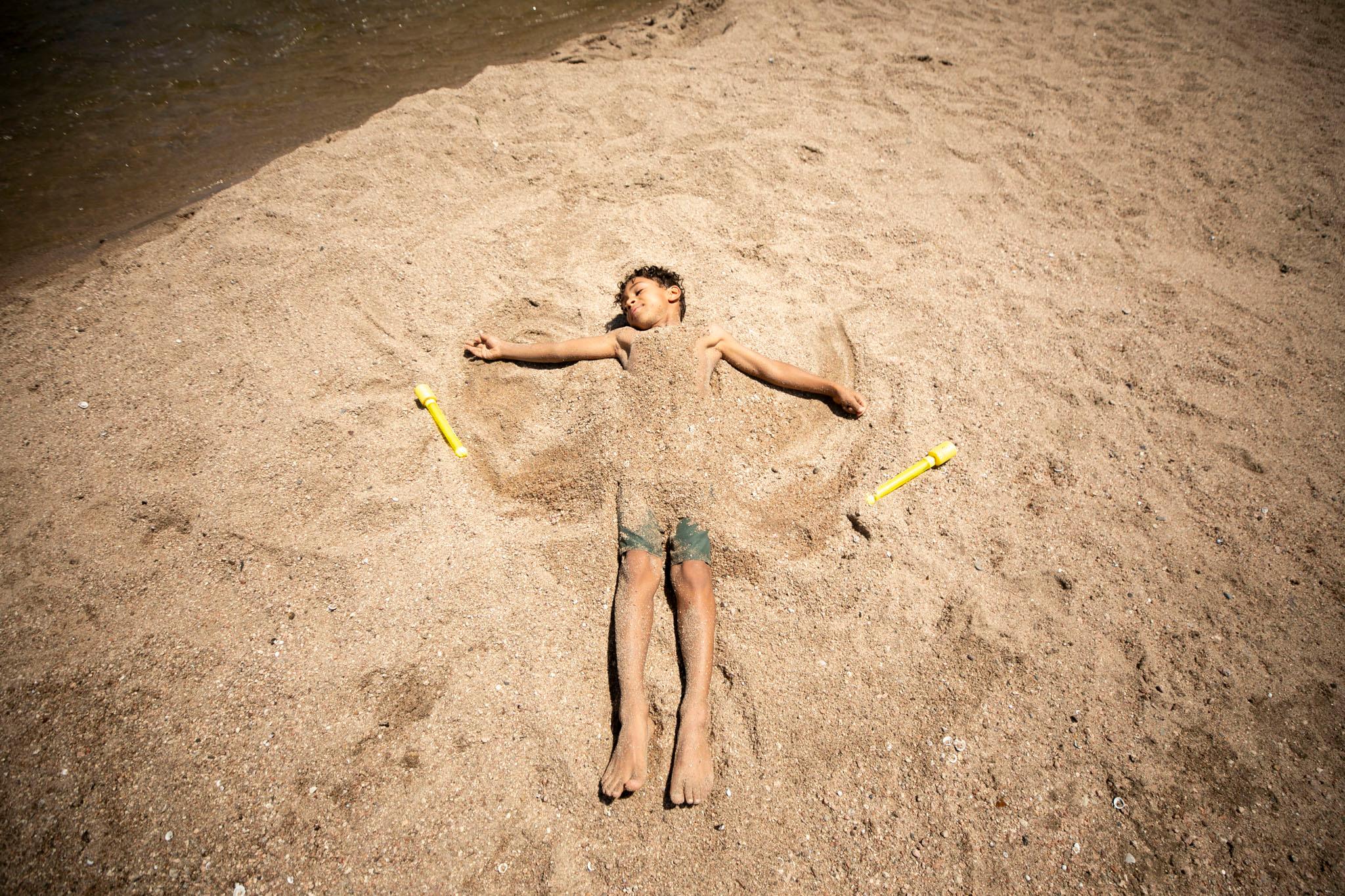 A kid lays in the sand, covered by the sand, with his arms stretched out as if it were winter and he was making a snow angel.