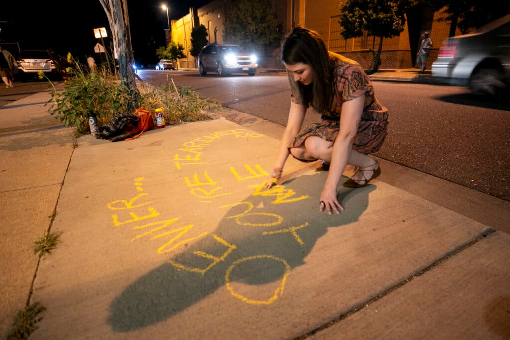 A woman in a dress leans over the sidewalk, writing in yellow chalk.