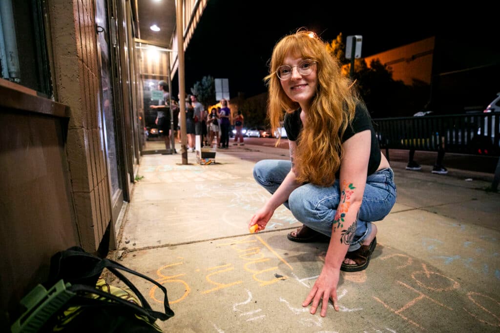 A woman in bangs and jeans squats on the sidewalk, which is covered with messages, with a piece of orange chalk in her hand. She smiles at the camera.