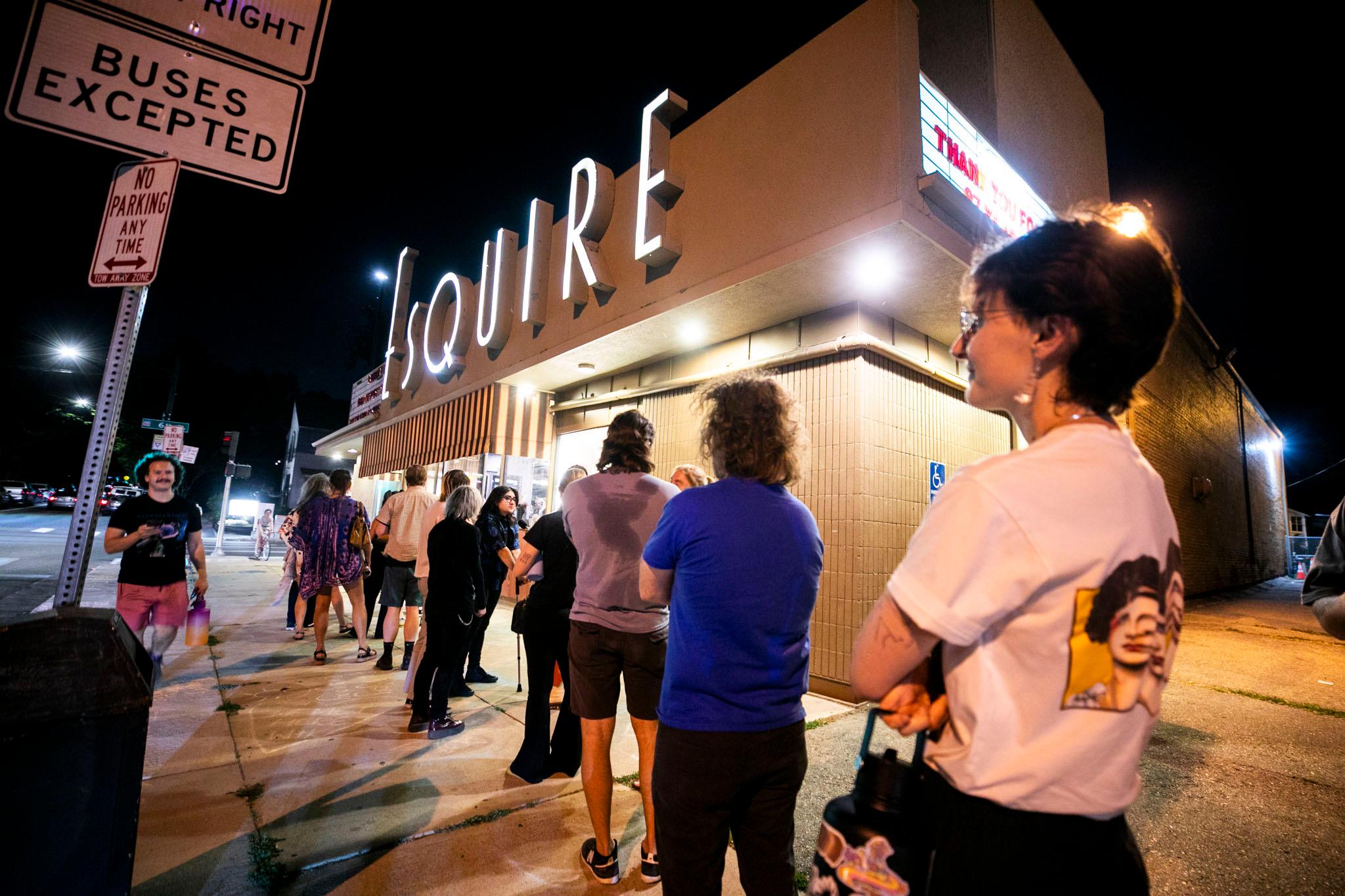 The Esquire Theater's illuminated letters glow above a lone of people waiting on the sidewalk to get inside.