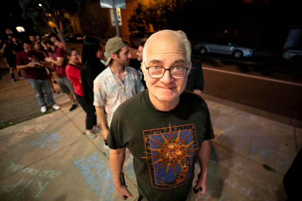 A man in glasses stares into the camera as he waits in a long line of people on the sidewalk; chalk messages cover the ground beneath him.