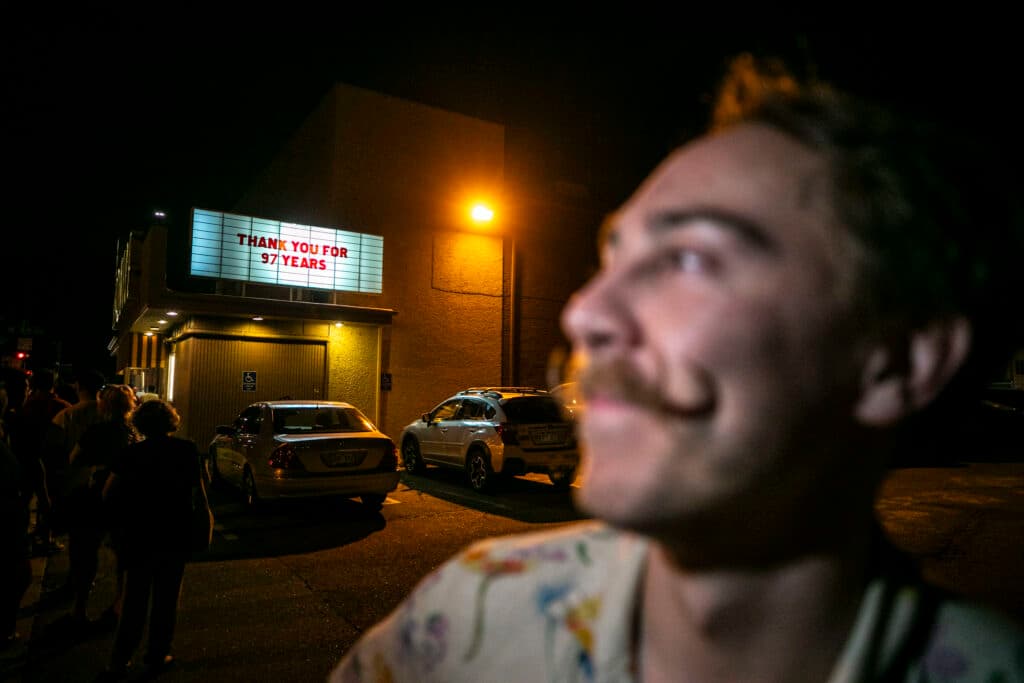 A mustachioed man is out of focus in the foreground; behind him a marquee reads "Thank you for 97 years."