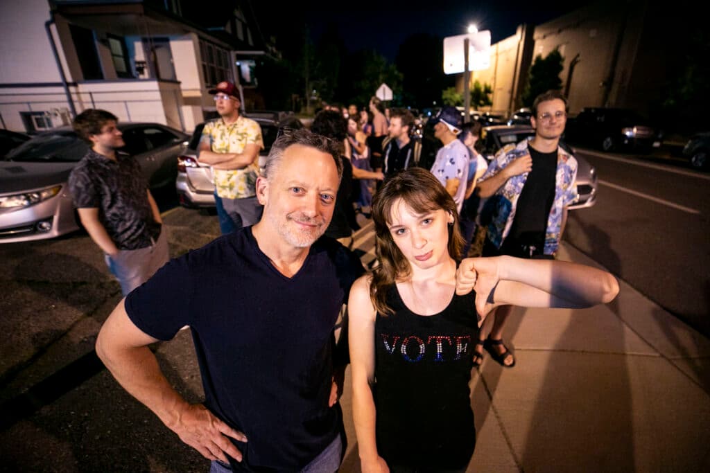 A man and a woman look right at the camera; she makes a thumbs-down gesture and puffs her lip out in a frown. They're in a big line of people on the sidewalk.