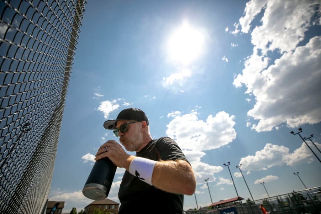 A man in a ballcap and shades holds a water bottle as his cheeks puff up with liquid. The sun shines bright above him.