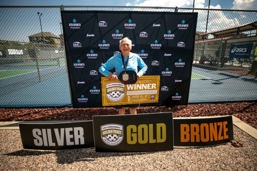 A man with white hair and a mostly bald head grins wide as he holds a sign that says &quot;WINNER,&quot; behind signs that say &quot;SILVER,&quot; &quot;GOLD&quot; and &quot;BRONZE.&quot;