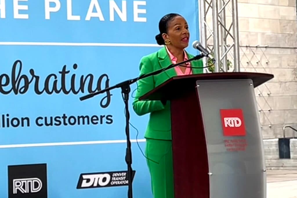 A woman in a shamrock green suit stands in front of a mic and a podium, which reads &quot;RTD.&quot;