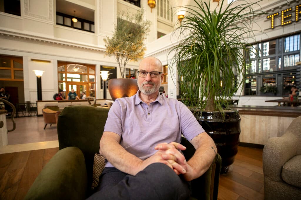 A man sits as if he's posing for a portrait for his first nonfiction book on sheepherding, a direct confident look into the camera, while sitting in a tall, well-lit room filled with plants and furniture.