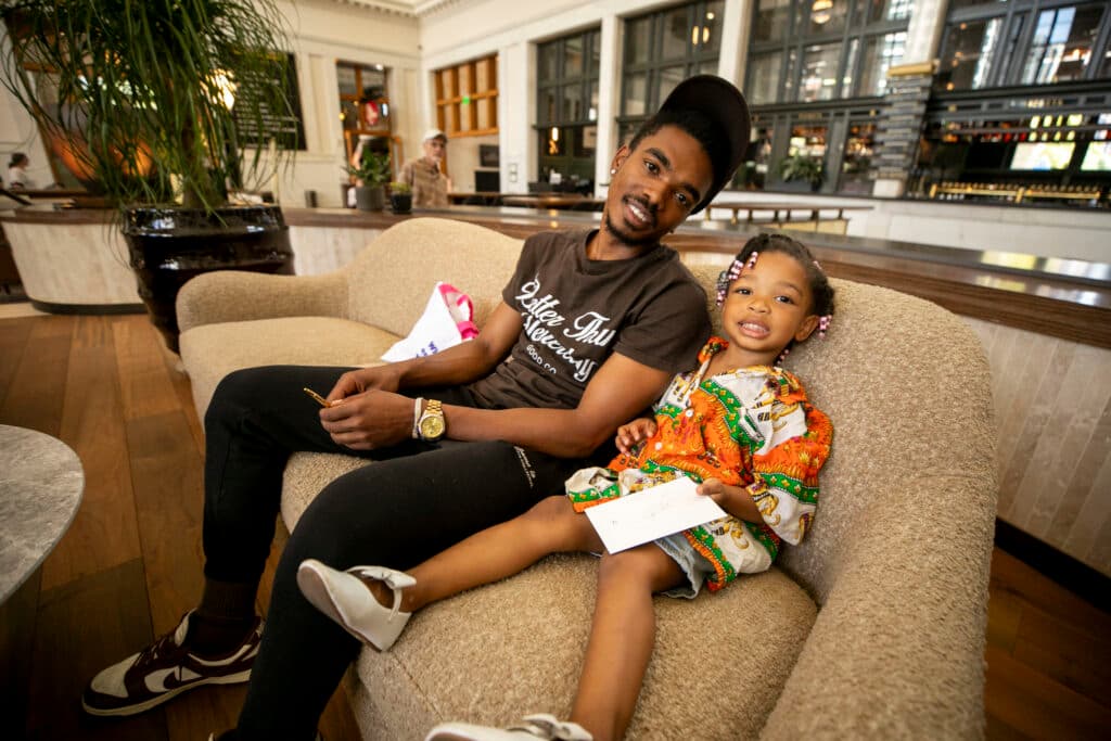 A man and a little girl smile at the camera from a beige couch in a big, well-lit room.