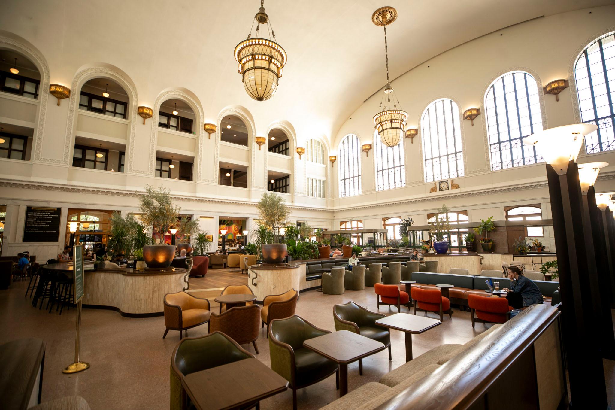 A well lit room with very tall ceilings is filled with earth-toned furniture and people sitting and working.