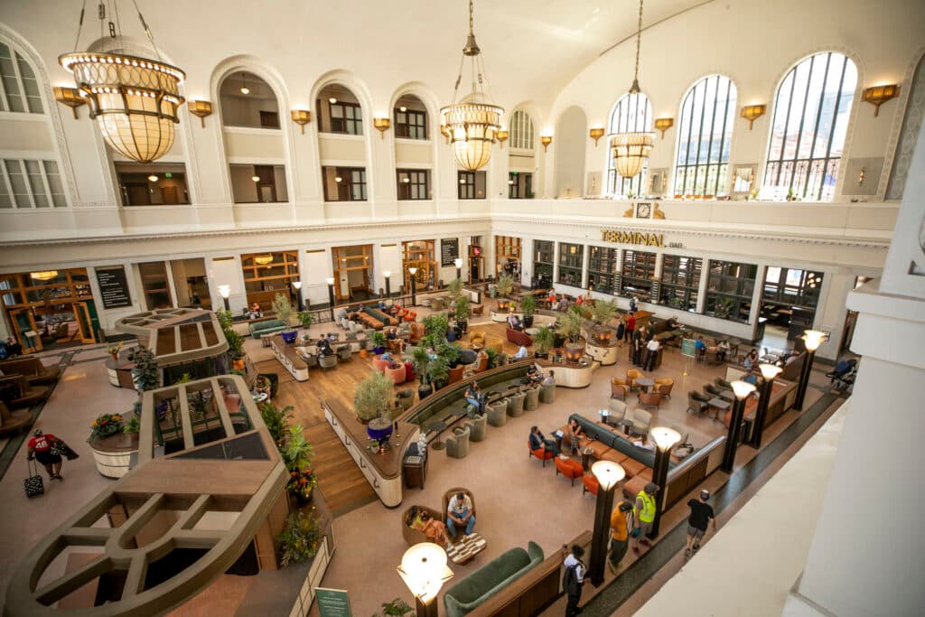 A well lit room, seen all at once from above, with very tall ceilings is filled with earth-toned furniture and people sitting and working.