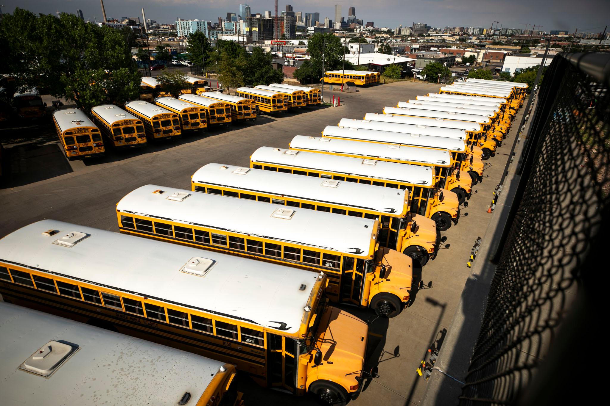 Many yellow school buses with white roofs, seen from above. The city skyline rises in the distance.