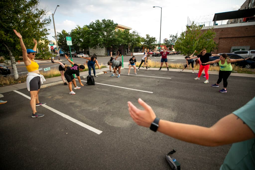 People in athletic gear wave their hands above their heads, stretching in a circle in a parking lot.