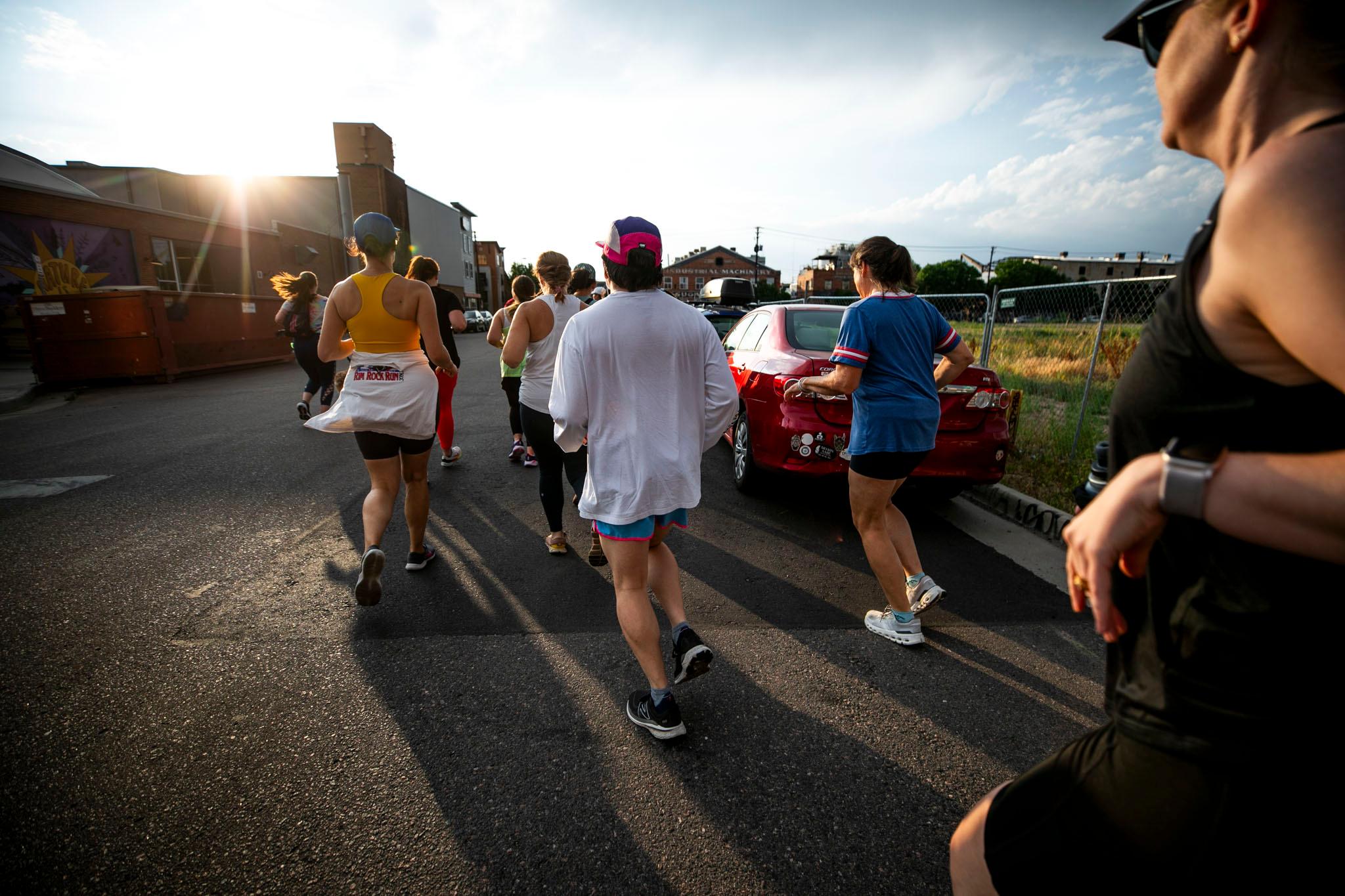 A group of people in athletic gear run towards the sun as it lowers towards the horizon, illuminated by early golden hour light.