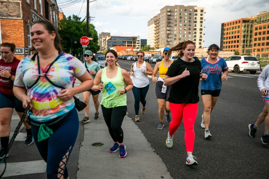 A group of people in athletic gear run down a sidewalk, smiling in stride.