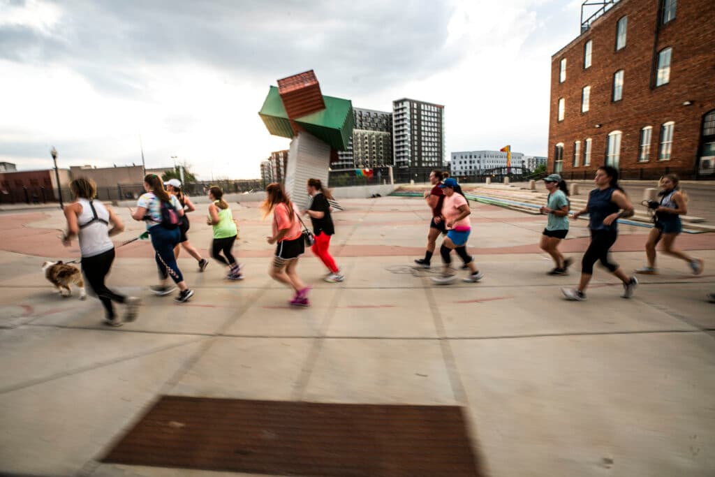 A group of people in athletic gear run through a plaza; their feet are blurred, the result of a slow shutter.