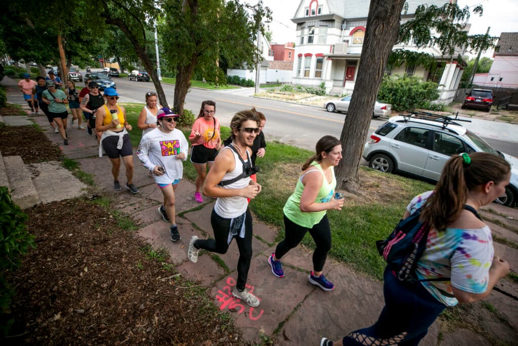 A group of people in athletic gear run over a sidewalk, smiling as they jog.