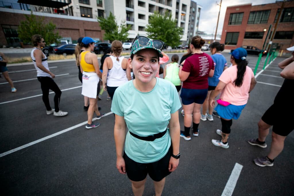 A woman in athletic gear smiles at the camera while standing in a parking lot.